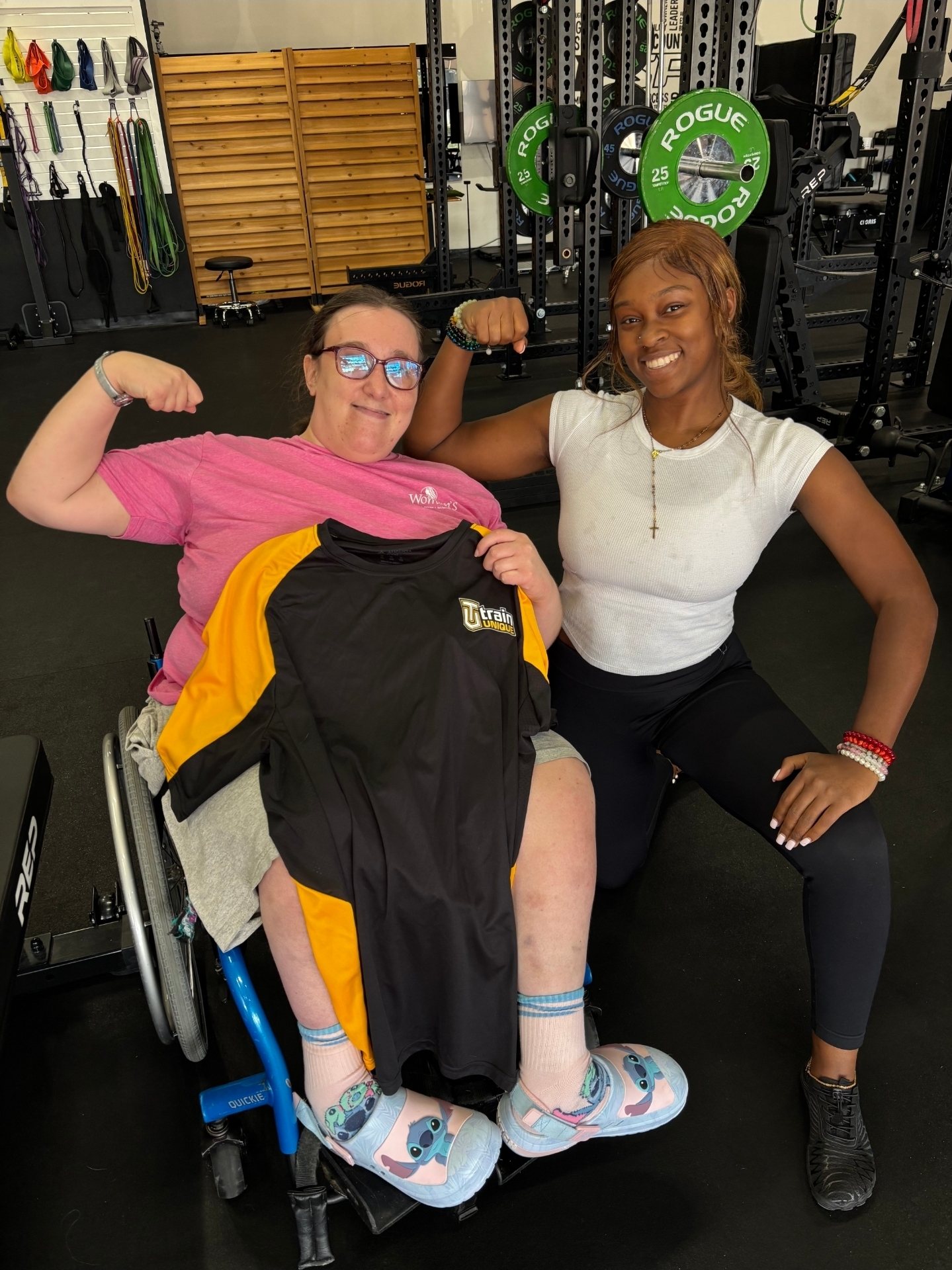 Dyimond kneeling and flexing beside one of her athletes, who is holding up her new Train Unique shirt from her wheelchair
