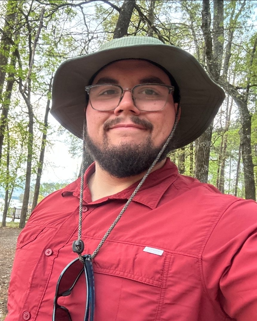 Coach Jessie in a sun hat and red shirt, trees and a lake behind him